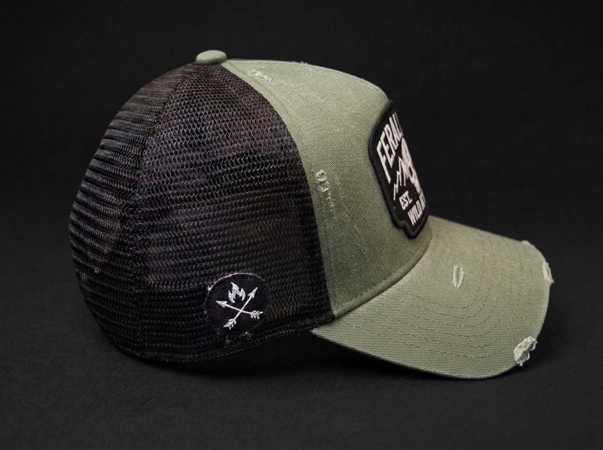 An olive green and black trucker cap with a distressed finish, featuring a black mesh back, a black embroidered patch on the side, and a black rectangular patch on the front brim.