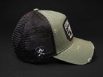 An olive green and black trucker cap with a distressed finish, featuring a black mesh back, a black embroidered patch on the side, and a black rectangular patch on the front brim.