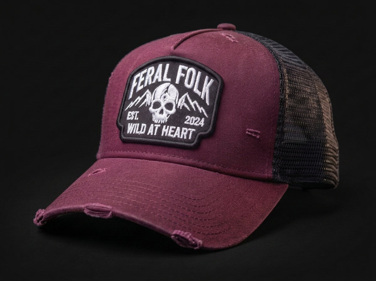 A distressed trucker cap in maroon with a black mesh back and a prominent black and white 'Feral Folk' skull patch on the front, set against a solid black background.