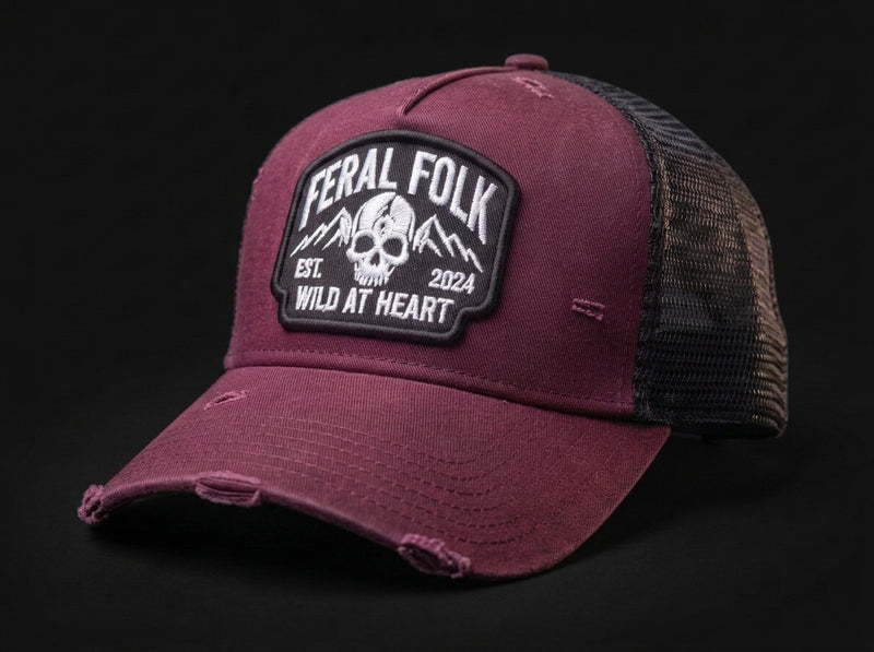 A distressed trucker cap in maroon with a black mesh back and a prominent black and white 'Feral Folk' skull patch on the front, set against a solid black background.