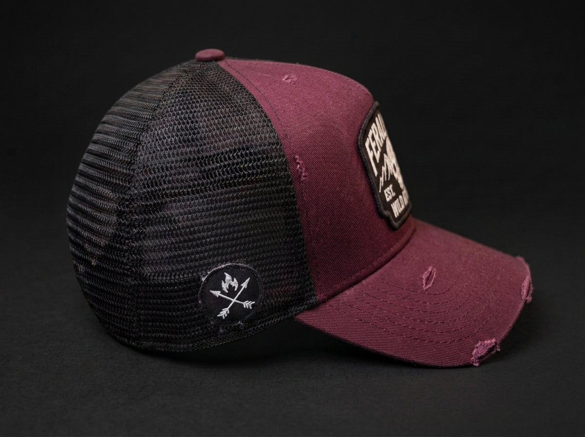 A distressed trucker cap in maroon and black mesh, featuring a 'Falcon' brand patch on the brim and a circular logo patch on the side, set against a dark background.