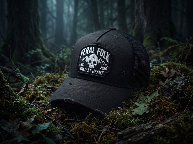 A black distressed trucker hat with a white embroidered 'Feral Folk' skull patch, resting on mossy forest floor logs in a dark, misty woodland setting.