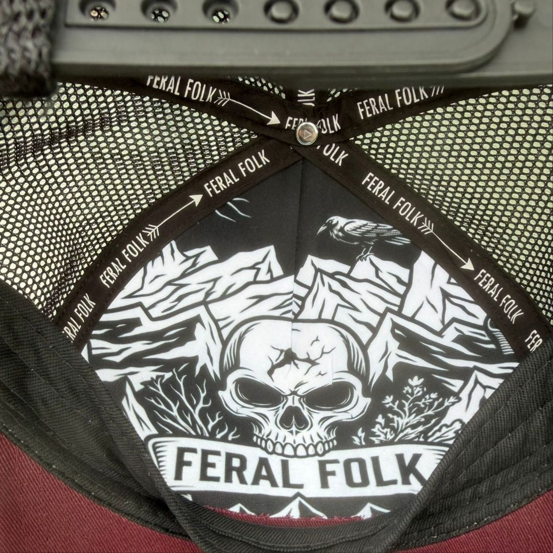 Close-up of the interior of a Feral Folk trucker hat, showing a black and white graphic print of a skull and mountain raven on the adjustable black mesh back, with the brand name 'FERAL FOLK' printed repeatedly on the elastic band.