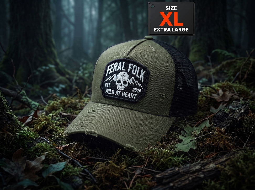 An olive green and black trucker hat with a distressed finish, featuring a 'Feral Folk' skull patch on the front. The hat is placed on a forest floor covered in moss and leaves, with a 'SIZE XL EXTRA LARGE' label overlay in the top right corner.