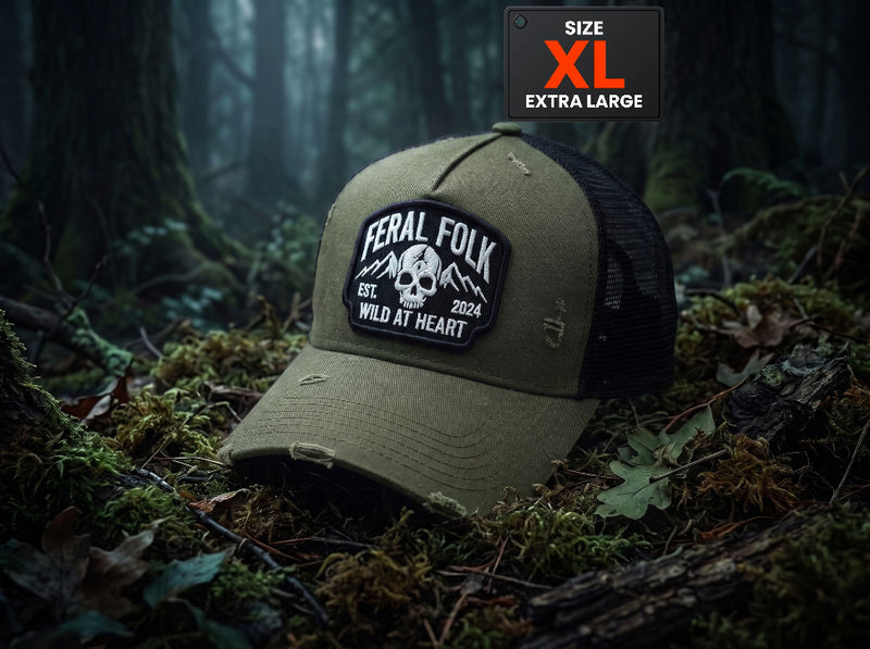 An olive green and black trucker hat with a distressed finish, featuring a 'Feral Folk' skull patch on the front. The hat is placed on a forest floor covered in moss and leaves, with a 'SIZE XL EXTRA LARGE' label overlay in the top right corner.