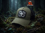 An olive green and black trucker hat with a distressed finish, featuring a 'Feral Folk' skull patch on the front. The hat is placed on a forest floor covered in moss and leaves, with a 'SIZE XL EXTRA LARGE' label overlay in the top right corner.