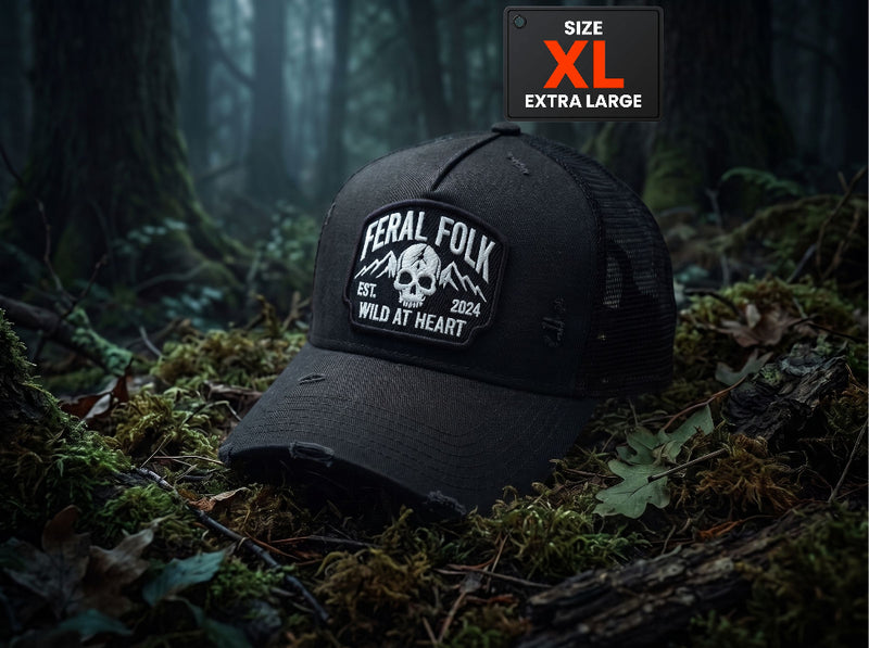 A black distressed trucker hat with a white embroidered 'Feral Folk' skull patch, resting on mossy forest floor logs. A black 'SIZE XL EXTRA LARGE' overlay badge is in the top right corner.