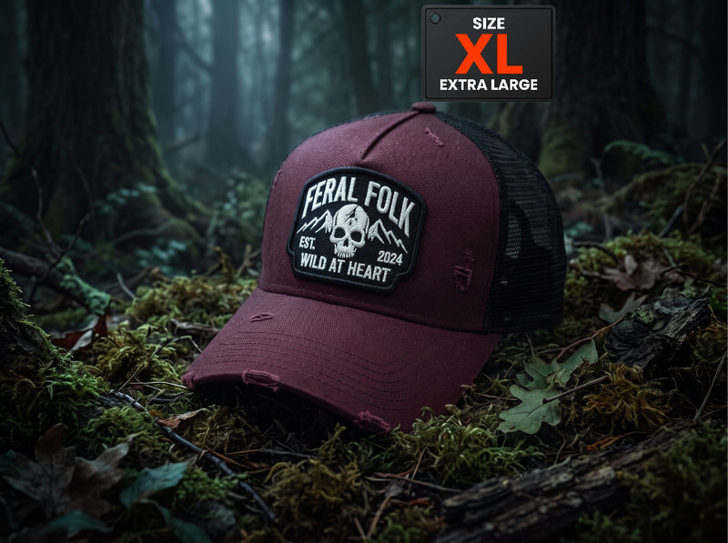 A distressed trucker hat in maroon and black mesh, placed on mossy forest floor with tree trunks in the background. The hat has a rectangular patch reading 'Feral Folk' with a skull graphic.