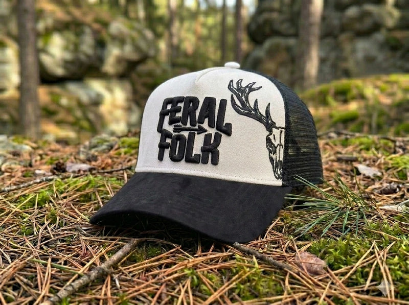 Black and white cap with 'Feral Folk' text and deer design on a forest floor.