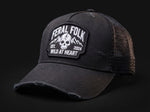 A black distressed trucker hat with a black mesh back, featuring a prominent embroidered patch on the front that reads 'FERAL FOLK' with a skull and mountain graphic and 'WILD AT HEART EST. 2024'.