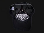 Black trucker-style cap shown from the back, featuring a black mesh back, black adjustable strap with white text, and a white graphic print of a skull and roses on the back panel.