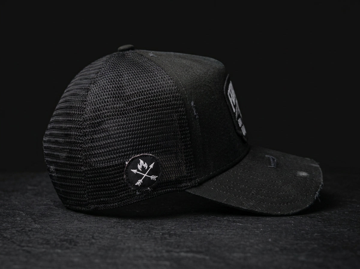 A black trucker-style cap with a mesh back and a curved brim, photographed on a dark textured surface against a black background. A circular patch with crossed arrows and a flame is visible on the side, and a rectangular patch appears on the brim.