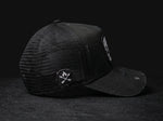A black trucker-style cap with a mesh back and a curved brim, photographed on a dark textured surface against a black background. A circular patch with crossed arrows and a flame is visible on the side, and a rectangular patch appears on the brim.