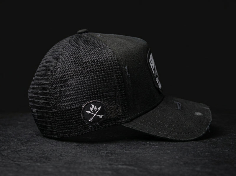 A black trucker-style cap with a mesh back and a curved brim, photographed on a dark textured surface against a black background. A circular patch with crossed arrows and a flame is visible on the side, and a rectangular patch appears on the brim.